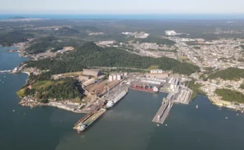 Portos de SC batem recorde com 65,7 milhões de toneladas Vista aérea de um grande porto industrial com navios cargueiros atracados, silos, armazéns e pátios de carga, situado ao lado de uma cidade costeira e morros arborizados sob um céu ensolarado.