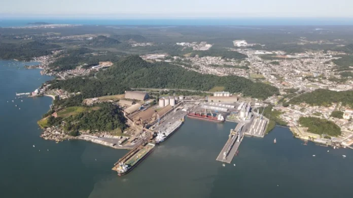 Portos de SC batem recorde com 65,7 milhões de toneladas Vista aérea de um grande porto industrial com navios cargueiros atracados, silos, armazéns e pátios de carga, situado ao lado de uma cidade costeira e morros arborizados sob um céu ensolarado.