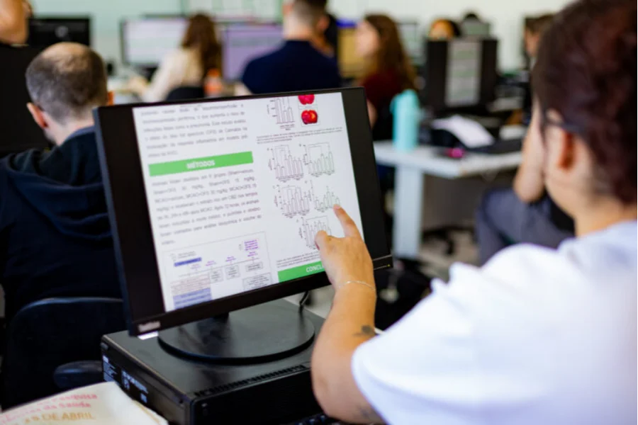Pesquisador em laboratório de informática da Unesc aponta para tela de computador exibindo gráficos e dados científicos, ilustrando o trabalho de pesquisa e a qualidade dos programas de pós-graduação da instituição, reconhecidos na avaliação da Capes.