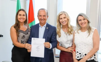Professora da Unesc assume nova diretoria da Educação em SC O governador Jorginho Mello, sorrindo, segura um documento oficial com a secretária Luciane Ceretta e a nova diretora Indianara Reynaud, acompanhadas pela reitora Gisele Coelho Lopes, todas sorridentes em um escritório com as bandeiras de Santa Catarina e do Brasil ao fundo.
