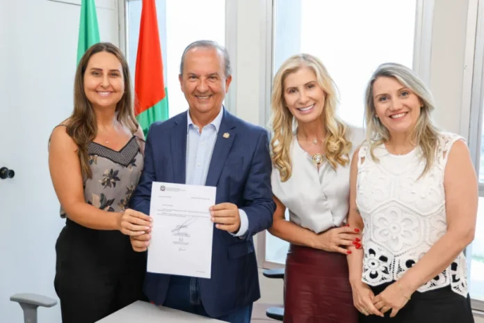 Professora da Unesc assume nova diretoria da Educação em SC O governador Jorginho Mello, sorrindo, segura um documento oficial com a secretária Luciane Ceretta e a nova diretora Indianara Reynaud, acompanhadas pela reitora Gisele Coelho Lopes, todas sorridentes em um escritório com as bandeiras de Santa Catarina e do Brasil ao fundo.