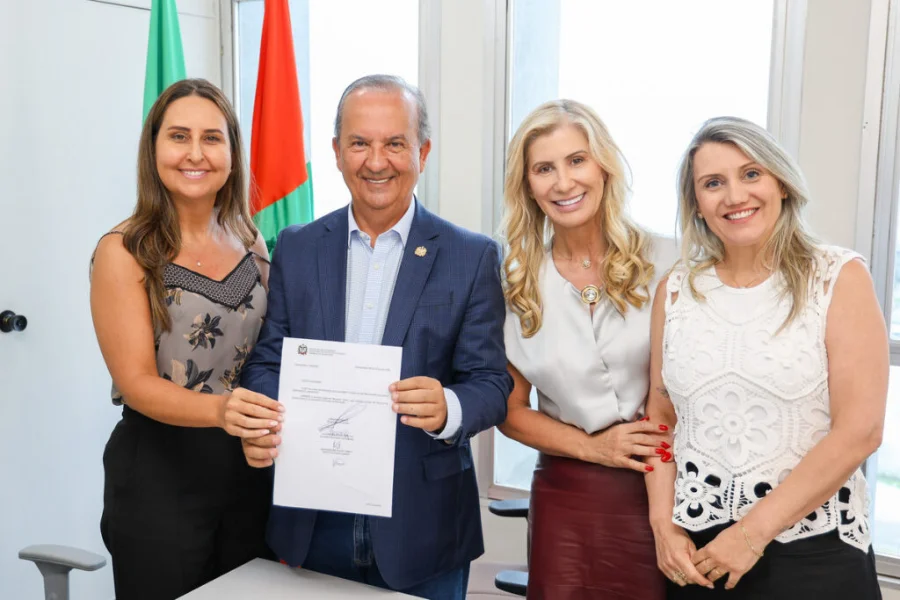 O governador Jorginho Mello, sorrindo, segura um documento oficial com a secretária Luciane Ceretta e a nova diretora Indianara Reynaud, acompanhadas pela reitora Gisele Coelho Lopes, todas sorridentes em um escritório com as bandeiras de Santa Catarina e do Brasil ao fundo.