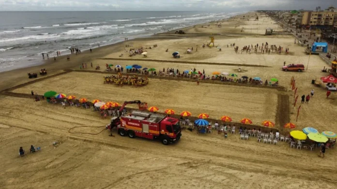 Programa Golfinho abre inscrições no Litoral Sul de SC Foto aérea mostra crianças reunidas na areia da praia para o Programa Golfinho do Corpo de Bombeiros. Na cena, um caminhão da corporação e banhistas sob guarda-sóis coloridos emolduram o grupo de alunos em uniformes amarelos. O mar e a orla do litoral catarinense aparecem ao fundo.