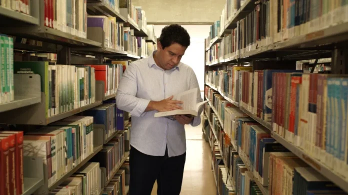 Programa Universidade Gratuita inscrições vão até dia 27 Estudante consultando um livro em uma biblioteca universitária repleta de estantes, ilustrando o acesso ao ensino superior proporcionado pelo programa Universidade Gratuita em Santa Catarina.