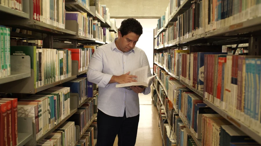 Estudante consultando um livro em uma biblioteca universitária repleta de estantes, ilustrando o acesso ao ensino superior proporcionado pelo programa Universidade Gratuita em Santa Catarina.