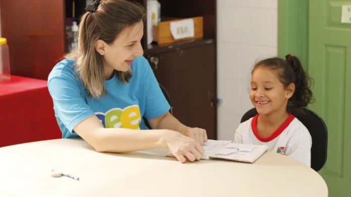 Rede Municipal de Criciúma inicia ano letivo com novidades Professora e aluna sorriem durante atividade pedagógica em sala de aula da rede municipal de Criciúma, representando o acolhimento e a preparação para o início do ano letivo de 2026.