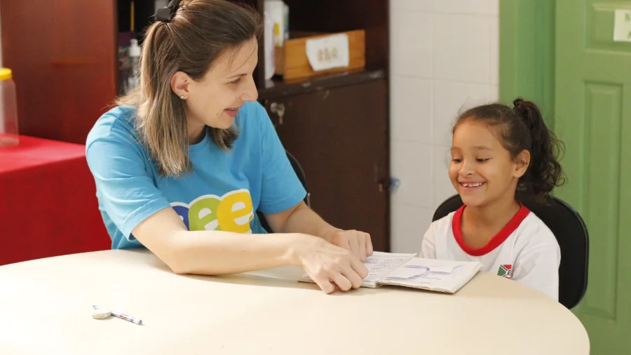 Professora e aluna sorriem durante atividade pedagógica em sala de aula da rede municipal de Criciúma, representando o acolhimento e a preparação para o início do ano letivo de 2026.