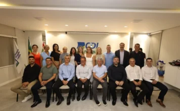 Rejane Silva de Araújo assume presidência da CDL Araranguá Foto de grupo com homens e mulheres, alguns sentados e outros em pé, posando sorridentes em uma sala bem iluminada. Ao fundo, uma parede branca exibe o logotipo azul e verde da "CDL Araranguá", ladeado por bandeiras do Brasil e de Santa Catarina, e um banner da Câmara de Dirigentes Lojistas à esquerda.