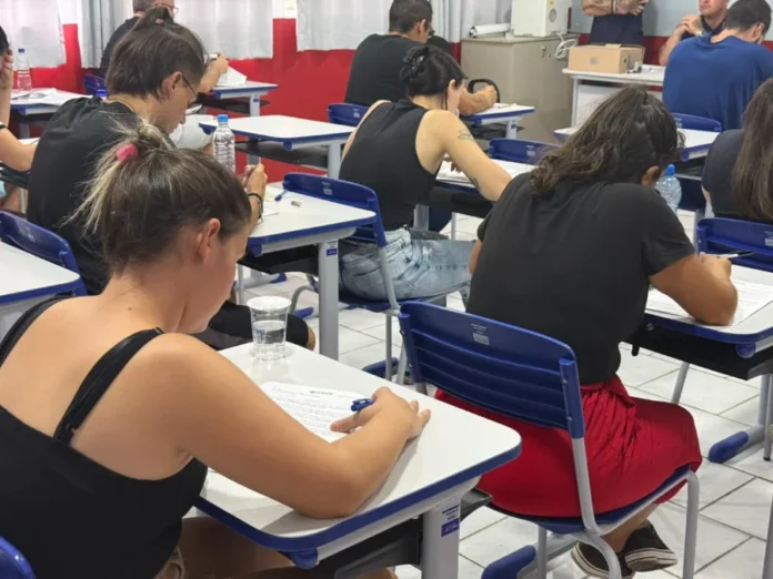 SAMAE Araranguá divulga classificação de processo seletivo Uma sala de aula com várias pessoas sentadas em carteiras escolares, vistas de costas, realizando a prova escrita do processo seletivo simplificado do SAMAE de Araranguá.