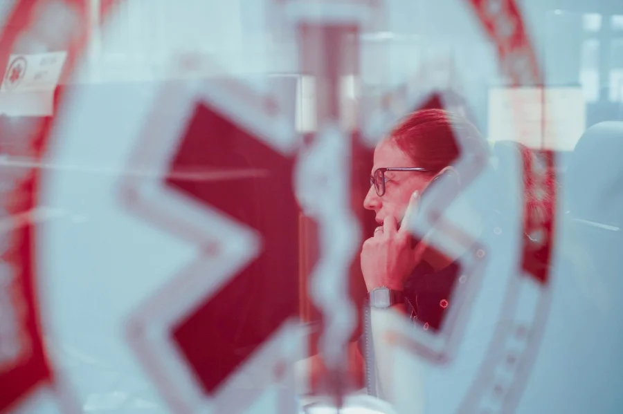 SAMU de SC registra queda de 4,6% nos trotes em 2025 Profissional do SAMU de Santa Catarina atendendo um chamado telefônico na central de regulação, vista através de um vidro com o logotipo do serviço. A imagem ilustra o trabalho de atendimento, que inclui a triagem de ligações e o combate aos trotes.