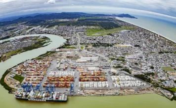 SC bate recorde histórico com exportações de US$ 12,2 bi Uma fotografia aérea em grande angular mostra um porto de contêineres movimentado na foz de um rio, cercado por uma cidade costeira e montanhas ao fundo. O porto está repleto de fileiras organizadas de contêineres coloridos em tons de vermelho, azul, amarelo e branco. No cais, vários guindastes pórticos azuis estão posicionados sobre um grande navio porta-contêineres da Evergreen, que está sendo carregado ou descarregado. A cidade se espalha ao redor do complexo portuário, com casas, prédios e estradas. Um aeroporto com pista pavimentada é visível na parte superior da imagem, próximo à costa e ao mar. As montanhas verdes se estendem ao longe sob um céu nublado. A água do rio e do mar tem uma tonalidade esverdeada.