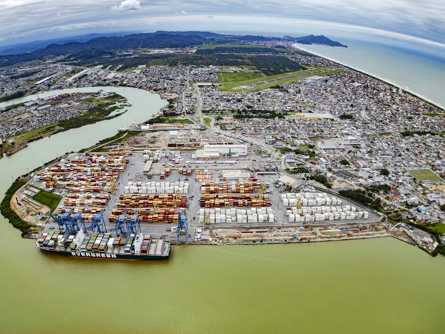 SC bate recorde histórico com exportações de US$ 12,2 bi Uma fotografia aérea em grande angular mostra um porto de contêineres movimentado na foz de um rio, cercado por uma cidade costeira e montanhas ao fundo. O porto está repleto de fileiras organizadas de contêineres coloridos em tons de vermelho, azul, amarelo e branco. No cais, vários guindastes pórticos azuis estão posicionados sobre um grande navio porta-contêineres da Evergreen, que está sendo carregado ou descarregado. A cidade se espalha ao redor do complexo portuário, com casas, prédios e estradas. Um aeroporto com pista pavimentada é visível na parte superior da imagem, próximo à costa e ao mar. As montanhas verdes se estendem ao longe sob um céu nublado. A água do rio e do mar tem uma tonalidade esverdeada.