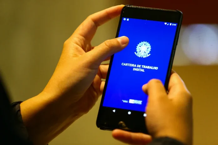 SC cria 59,2 mil empregos em 2025 e indica desaceleração Foto em close de mãos segurando um smartphone que exibe a tela inicial do aplicativo da Carteira de Trabalho Digital, com o brasão da República sobre um fundo azul. A imagem simboliza a formalização de novos postos de trabalho em Santa Catarina.
