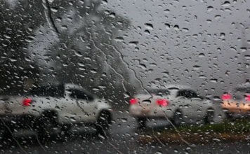 SC em alerta máximo para temporais nesta quinta-feira Visão de dentro de um carro através de um para-brisa coberto de gotas de chuva, mostrando veículos com luzes traseiras vermelhas acesas em uma estrada molhada e escura. A imagem ilustra as condições de tempo chuvoso e alerta para motoristas durante os temporais previstos em Santa Catarina.