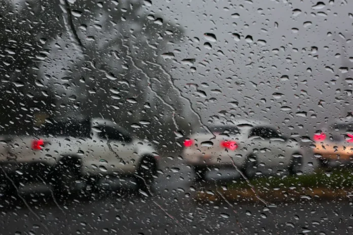 SC em alerta máximo para temporais nesta quinta-feira Visão de dentro de um carro através de um para-brisa coberto de gotas de chuva, mostrando veículos com luzes traseiras vermelhas acesas em uma estrada molhada e escura. A imagem ilustra as condições de tempo chuvoso e alerta para motoristas durante os temporais previstos em Santa Catarina.