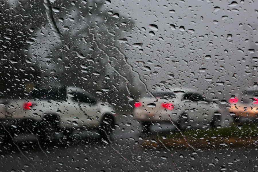 Visão de dentro de um carro através de um para-brisa coberto de gotas de chuva, mostrando veículos com luzes traseiras vermelhas acesas em uma estrada molhada e escura. A imagem ilustra as condições de tempo chuvoso e alerta para motoristas durante os temporais previstos em Santa Catarina.