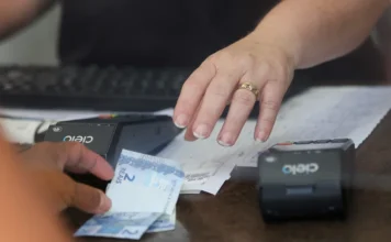 SC salta para 6º em ranking de liberdade econômica Close de mãos sobre um balcão realizando uma transação comercial com notas de dinheiro e uma máquina de cartão, ilustrando o movimento da economia e o fortalecimento do ambiente de negócios em Santa Catarina.