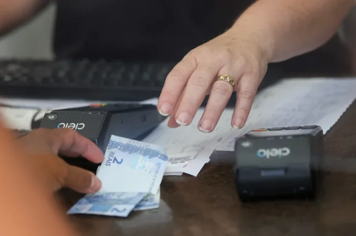 SC salta para 6º em ranking de liberdade econômica Close de mãos sobre um balcão realizando uma transação comercial com notas de dinheiro e uma máquina de cartão, ilustrando o movimento da economia e o fortalecimento do ambiente de negócios em Santa Catarina.