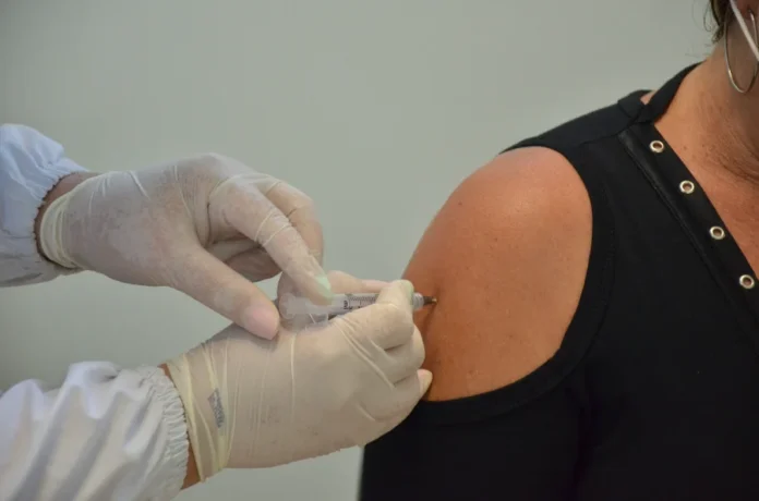 SC supera média nacional em 18 das 20 vacinas em 2025 Fotografia em close-up mostra as mãos de um profissional de saúde, utilizando luvas brancas de procedimento, no momento exato da aplicação de uma vacina no braço de uma pessoa. A imagem destaca a agulha sendo inserida no músculo deltoide, simbolizando o esforço contínuo das equipes de saúde de Santa Catarina para manter a população protegida contra doenças evitáveis.