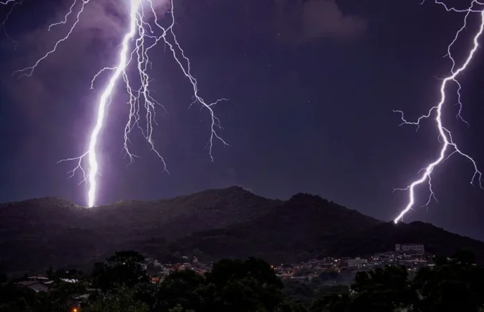 SC terá calor de 37°C e alerta para temporais na semana Fotografia noturna de raios potentes atingindo o topo de montanhas cobertas por vegetação, com uma área urbana iluminada na base. O céu está escuro e carregado, ilustrando o cenário de temporais isolados e instabilidade atmosférica previstos pela Defesa Civil de Santa Catarina.