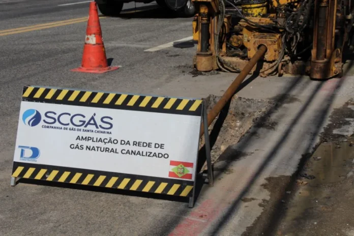 SCGÁS investirá R$ 104,8 milhões em Santa Catarina em 2026 Canteiro de obras em via pública com uma placa de sinalização da SCGÁS indicando "Ampliação da rede de gás natural canalizado". Ao fundo, um equipamento de perfuração insere a tubulação no solo, ilustrando os investimentos em infraestrutura urbana mencionados na matéria.