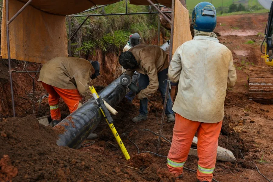 Trabalhadores com uniformes de segurança e equipamentos de proteção realizam a instalação e soldagem de uma tubulação de gás natural de grande porte em uma vala. A cena ilustra as obras de expansão da infraestrutura da SCGÁS em solo catarinense.