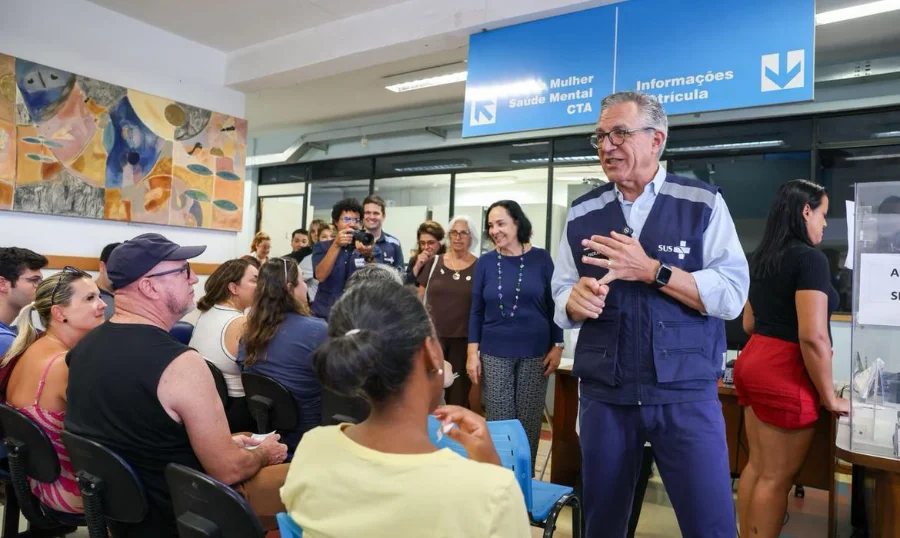 Ministro da Saúde, Alexandre Padilha, usando colete do SUS, fala com público em unidade de saúde durante anúncio sobre a vacinação contra a dengue.