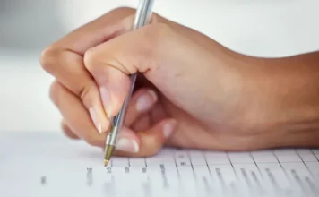 Saiu o gabarito do concurso da Saúde SC; veja como acessar Mão segurando uma caneta e preenchendo uma folha de respostas ou formulário, em alusão à realização das provas do concurso público da Secretaria de Estado da Saúde (SES).