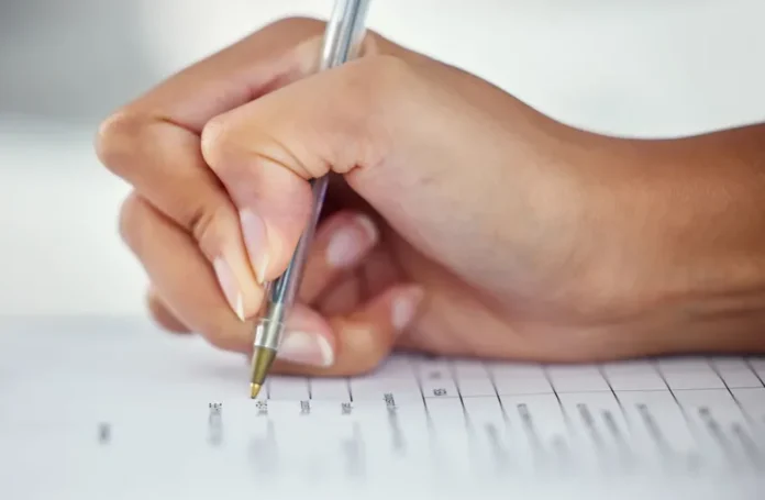 Saiu o gabarito do concurso da Saúde SC; veja como acessar Mão segurando uma caneta e preenchendo uma folha de respostas ou formulário, em alusão à realização das provas do concurso público da Secretaria de Estado da Saúde (SES).