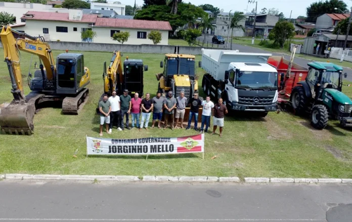 Santa Rosa do Sul renova frota com sete novos veículos Fotografia do prefeito de Santa Rosa do Sul, Almides Roberg da Rosa, e lideranças locais em frente à nova frota de máquinas pesadas. O grupo posa atrás de uma faixa com os dizeres "Obrigado Governador Jorginho Mello". Ao fundo, estão alinhadas duas escavadeiras de esteira, uma retroescavadeira, um caminhão caçamba branco e um trator agrícola verde.