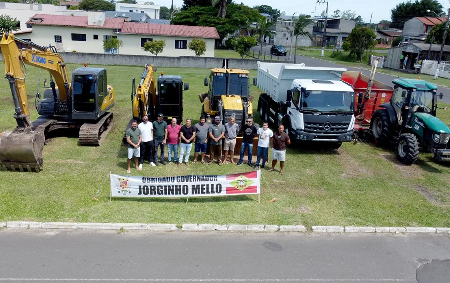 Fotografia do prefeito de Santa Rosa do Sul, Almides Roberg da Rosa, e lideranças locais em frente à nova frota de máquinas pesadas. O grupo posa atrás de uma faixa com os dizeres 