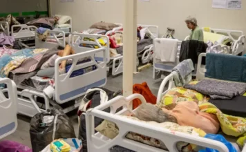 Saúde lança nova Caderneta da Pessoa Idosa com QR Codes Fotografia de um abrigo ou enfermaria com várias camas hospitalares brancas próximas umas das outras. Alguns idosos estão deitados sob cobertas coloridas, enquanto uma mulher idosa de blusa verde está sentada na beira de uma das camas. Sacolas e pertences pessoais aparecem no chão entre os leitos. A imagem ilustra a necessidade de acompanhamento e cuidado integral com a saúde da pessoa idosa.