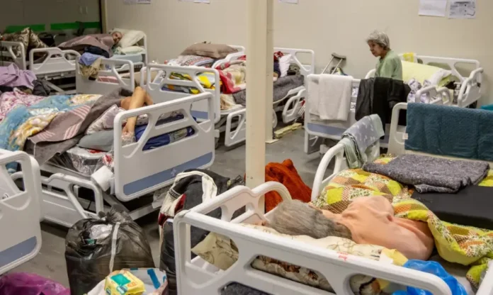 Saúde lança nova Caderneta da Pessoa Idosa com QR Codes Fotografia de um abrigo ou enfermaria com várias camas hospitalares brancas próximas umas das outras. Alguns idosos estão deitados sob cobertas coloridas, enquanto uma mulher idosa de blusa verde está sentada na beira de uma das camas. Sacolas e pertences pessoais aparecem no chão entre os leitos. A imagem ilustra a necessidade de acompanhamento e cuidado integral com a saúde da pessoa idosa.