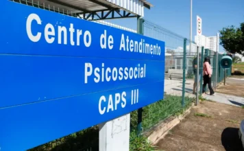 Saúde revisa normas e financiamento da rede psicossocial Placa azul de identificação de um Centro de Atendimento Psicossocial (CAPS III) em primeiro plano. Ao fundo, vê-se a estrutura externa da unidade de saúde sob um céu claro. A imagem ilustra a Rede de Atenção Psicossocial (Raps), cujas normas de financiamento e diretrizes estão sendo revisadas pelo Ministério da Saúde.