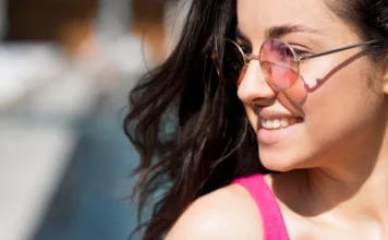 Saúde visual: Saiba como proteger os olhos no verão Close-up lateral de uma jovem sorrindo sob o sol. Ela possui cabelos escuros e usa óculos de sol com lentes arredondadas de cor rosada e armação metálica fina. A luz solar reflete em sua pele, destacando a importância da proteção ocular no verão.