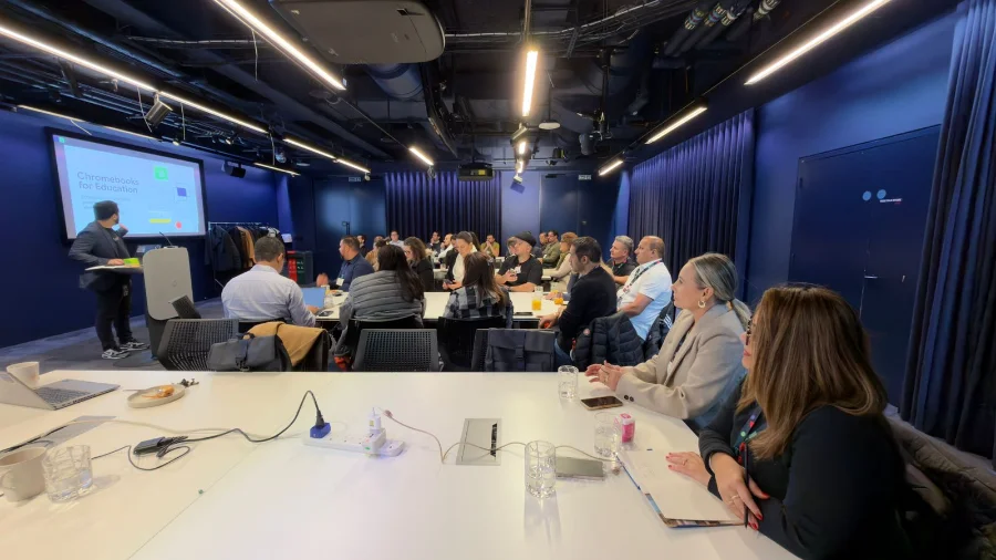 Foto interna de uma sala de conferência durante visita técnica à sede da Google em Londres. À direita, em primeiro plano, aparece a Secretária de Educação de Araranguá, Mariluce Rodrigues, sentada à mesa e olhando para uma apresentação projetada à frente. Ao fundo, um palestrante apresenta o tema 'Chromebooks for Education' para a delegação brasileira.