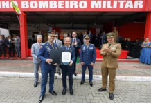 Segurança em SC recebe aporte de R$ 120 milhões em veículos Governador Jorginho Mello, ao centro, segura uma placa simbólica ladeado por oficiais do Corpo de Bombeiros Militar e da Polícia Militar durante solenidade em Florianópolis. O documento em suas mãos exibe o número "56" e o valor "R$ 76.610.753,26", representando a autorização para a compra da nova frota de caminhões anunciada na matéria.