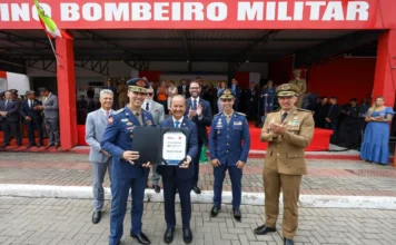 Segurança em SC recebe aporte de R$ 120 milhões em veículos Governador Jorginho Mello, ao centro, segura uma placa simbólica ladeado por oficiais do Corpo de Bombeiros Militar e da Polícia Militar durante solenidade em Florianópolis. O documento em suas mãos exibe o número "56" e o valor "R$ 76.610.753,26", representando a autorização para a compra da nova frota de caminhões anunciada na matéria.