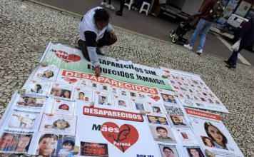 Senado analisa criação de delegacias de desaparecidos Mulher ajoelhada em uma calçada de mosaico português ao lado de banners e diversos cartazes espalhados com fotos e nomes de pessoas desaparecidas. Um dos banners centrais exibe o logotipo do grupo Mães do Paraná — um coração vermelho com a silhueta de um rosto feminino — acompanhado da frase "Desaparecidos sim. Esquecidos jamais!". O registro ilustra a mobilização social que fundamentou a criação do projeto de lei para delegacias especializadas.