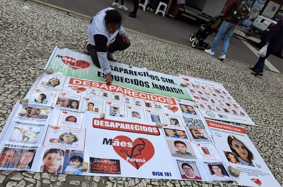 Mulher ajoelhada em uma calçada de mosaico português ao lado de banners e diversos cartazes espalhados com fotos e nomes de pessoas desaparecidas. Um dos banners centrais exibe o logotipo do grupo Mães do Paraná — um coração vermelho com a silhueta de um rosto feminino — acompanhado da frase 