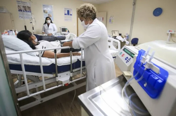 Senado analisa projeto com metas para Dia da Diálise Profissional de saúde supervisiona paciente durante sessão de diálise em ambiente hospitalar, com equipamento médico ao lado.