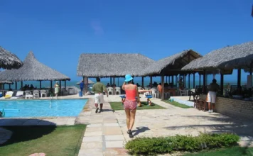 Senado avalia cobrança proporcional de diárias em hotéis Área de lazer com piscina e quiosques de palha em um hotel no Ceará, com pessoas caminhando em um dia ensolarado. A imagem ilustra um empreendimento hoteleiro que seria impactado pelo projeto que estabelece a cobrança proporcional da diária pelo tempo de permanência do hóspede.