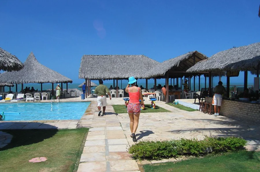 Área de lazer com piscina e quiosques de palha em um hotel no Ceará, com pessoas caminhando em um dia ensolarado. A imagem ilustra um empreendimento hoteleiro que seria impactado pelo projeto que estabelece a cobrança proporcional da diária pelo tempo de permanência do hóspede.