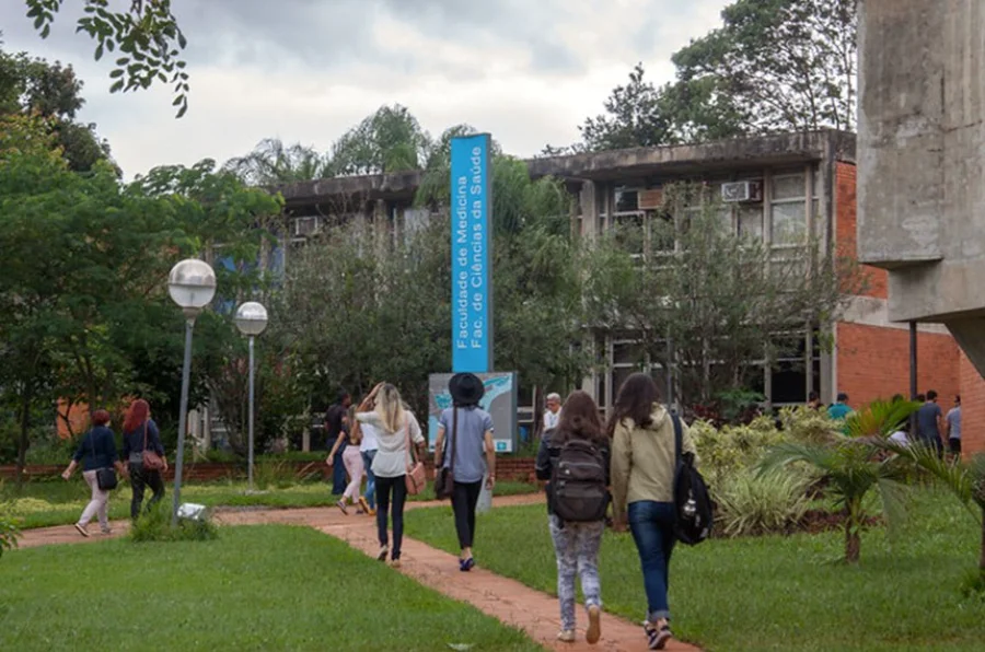 Estudantes caminham pelo campus em direção à entrada de um prédio identificado com uma placa azul onde se lê 