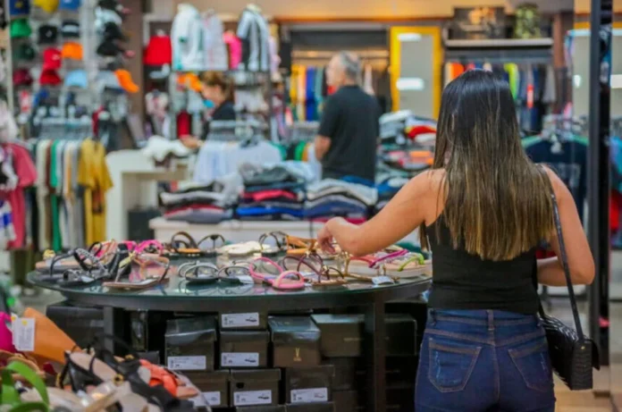 Senado avalia jornada de 36 horas e fim da reeleição Fotografia do interior de uma loja de roupas e calçados, mostrando uma cliente em primeiro plano observando sandálias em uma mesa de exposição e funcionários ao fundo. A imagem ilustra o setor de comércio e serviços, um dos segmentos mais impactados pela proposta de redução da jornada de trabalho para 36 horas semanais e a implementação da escala 5x2.
