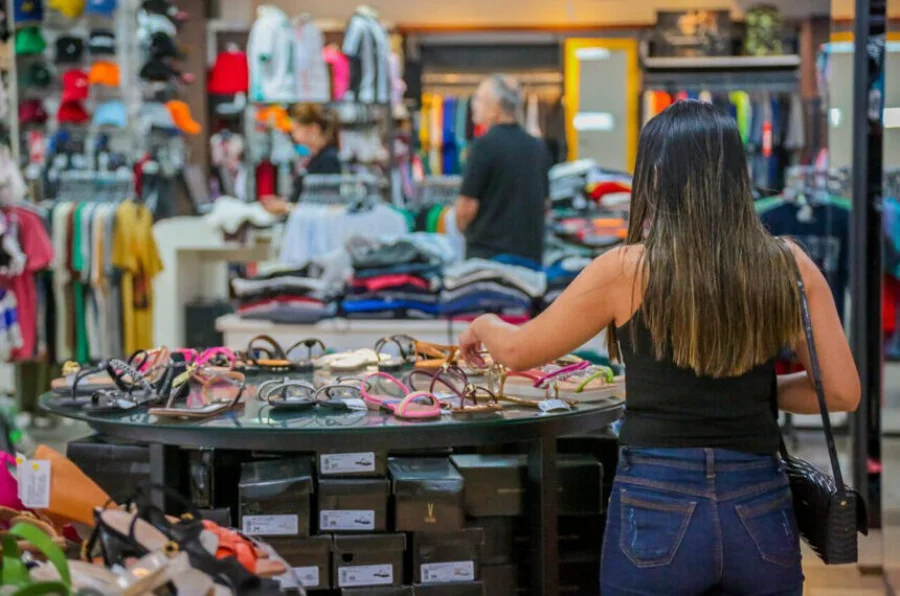 Fotografia do interior de uma loja de roupas e calçados, mostrando uma cliente em primeiro plano observando sandálias em uma mesa de exposição e funcionários ao fundo. A imagem ilustra o setor de comércio e serviços, um dos segmentos mais impactados pela proposta de redução da jornada de trabalho para 36 horas semanais e a implementação da escala 5x2.