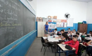 Senado prioriza novo Plano Nacional de Educação para 2026 Fotografia de uma sala de aula em escola pública com paredes brancas e ventilador. Uma professora está em pé junto ao quadro-negro observando alunos sentados em fileiras, que escrevem em seus cadernos. Ilustra a prioridade do Senado na votação do novo Plano Nacional de Educação.