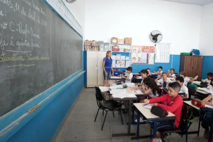 Senado prioriza novo Plano Nacional de Educação para 2026 Fotografia de uma sala de aula em escola pública com paredes brancas e ventilador. Uma professora está em pé junto ao quadro-negro observando alunos sentados em fileiras, que escrevem em seus cadernos. Ilustra a prioridade do Senado na votação do novo Plano Nacional de Educação.