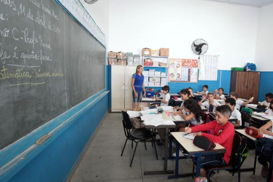 Senado prioriza novo Plano Nacional de Educação para 2026 Fotografia de uma sala de aula em escola pública com paredes brancas e ventilador. Uma professora está em pé junto ao quadro-negro observando alunos sentados em fileiras, que escrevem em seus cadernos. Ilustra a prioridade do Senado na votação do novo Plano Nacional de Educação.