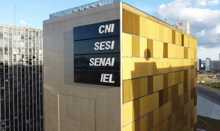 Setor produtivo e sindicatos criticam manutenção da Selic A imagem mostra a fachada do prédio da Confederação Nacional da Indústria (CNI), em Brasília. O edifício, de cor amarela, exibe as siglas CNI, SESI, SENAI e IEL em letras pretas. A CNI é uma das principais entidades que manifestaram forte oposição à manutenção da taxa Selic em 15% pelo Comitê de Política Monetária (Copom), argumentando que o nível elevado dos juros prejudica o setor produtivo e a geração de empregos.